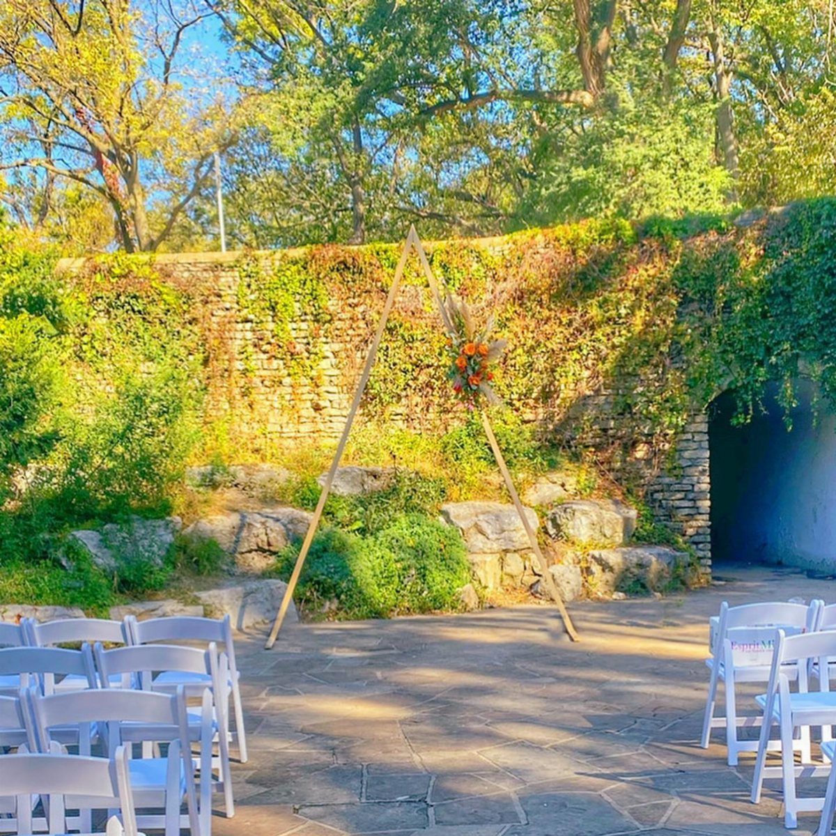 Wooden triangle wedding arch with floral arrangements against an ivy-covered stone wall.