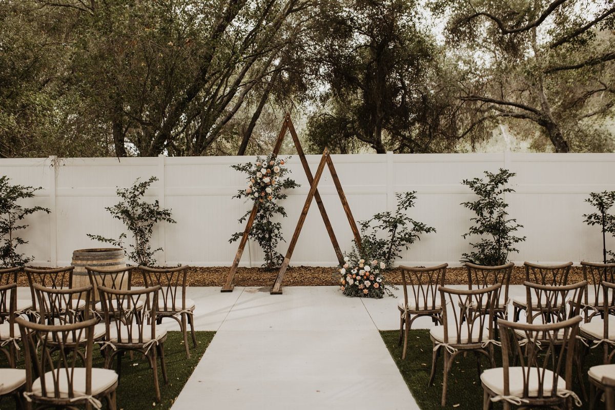 Wooden triangle wedding arch with minimal white and peach floral accents outdoors.