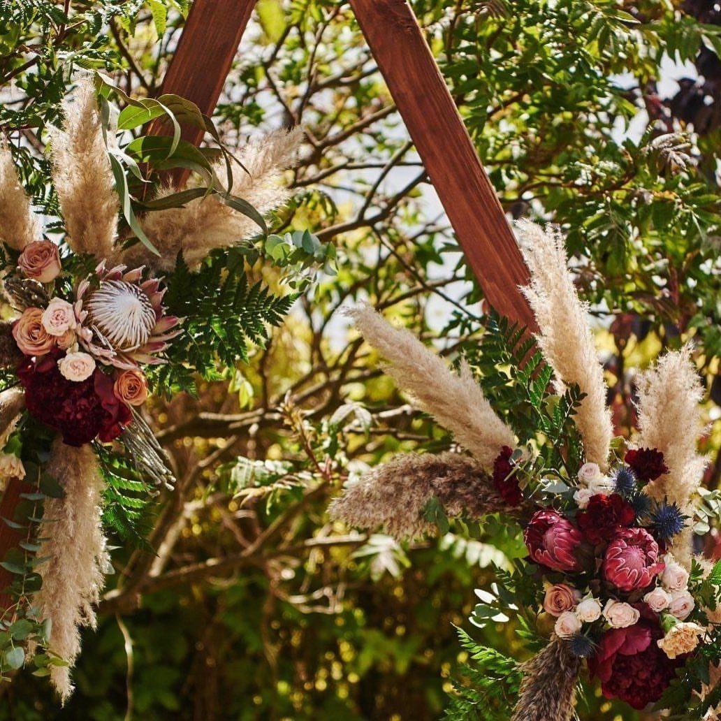 Wooden triangle wedding arch adorned with pampas grass and red floral arrangements outdoors.