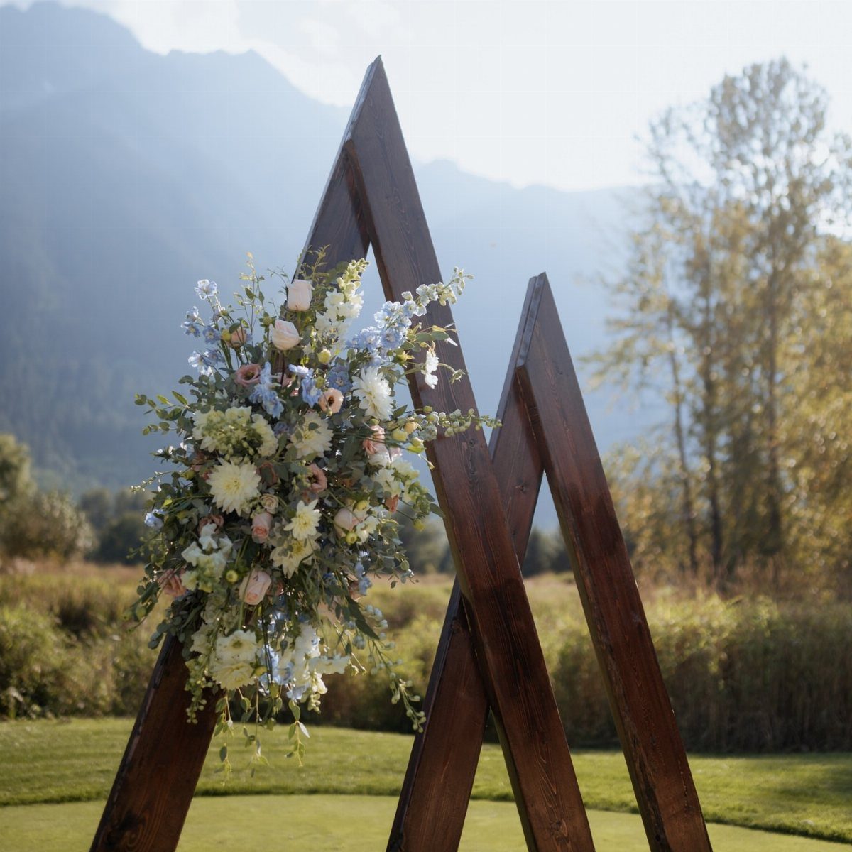 Wooden triangle wedding arch decorated with pastel flowers in an outdoor mountain setting.