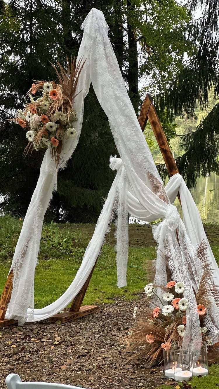 Wooden triangle wedding arch draped in white lace with floral arrangements outdoors.