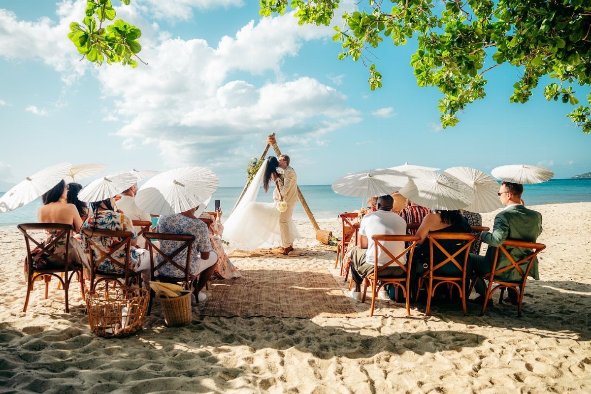 Couple marrying under a wooden triangle arch on a sunny beach with guests.