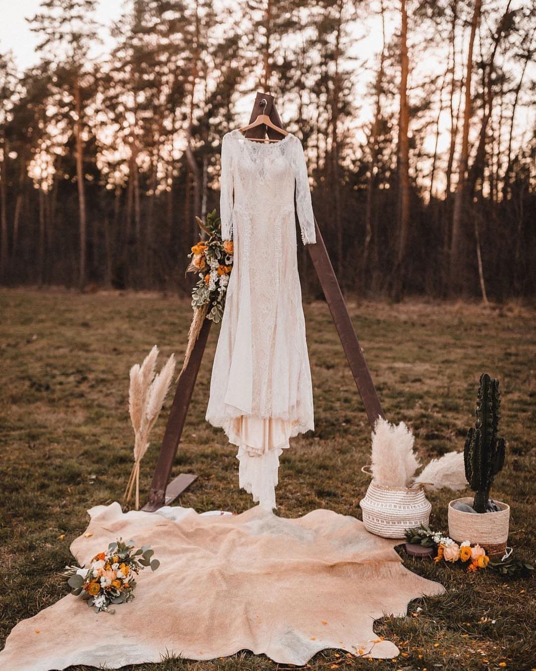 White wedding dress hanging on a wooden triangle arch in a forest field.