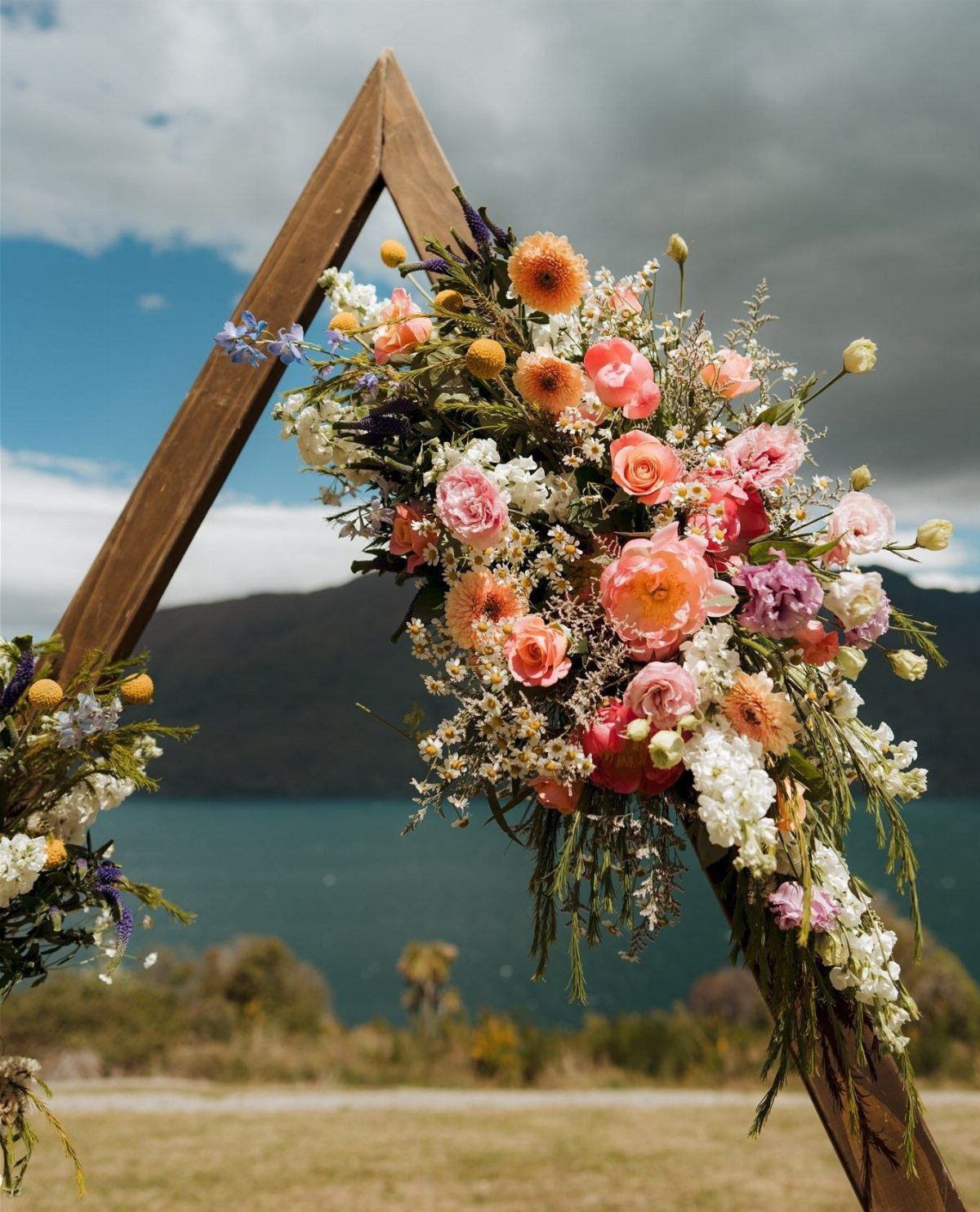 Wooden triangle wedding arch adorned with peach and pink floral arrangements by a lake.