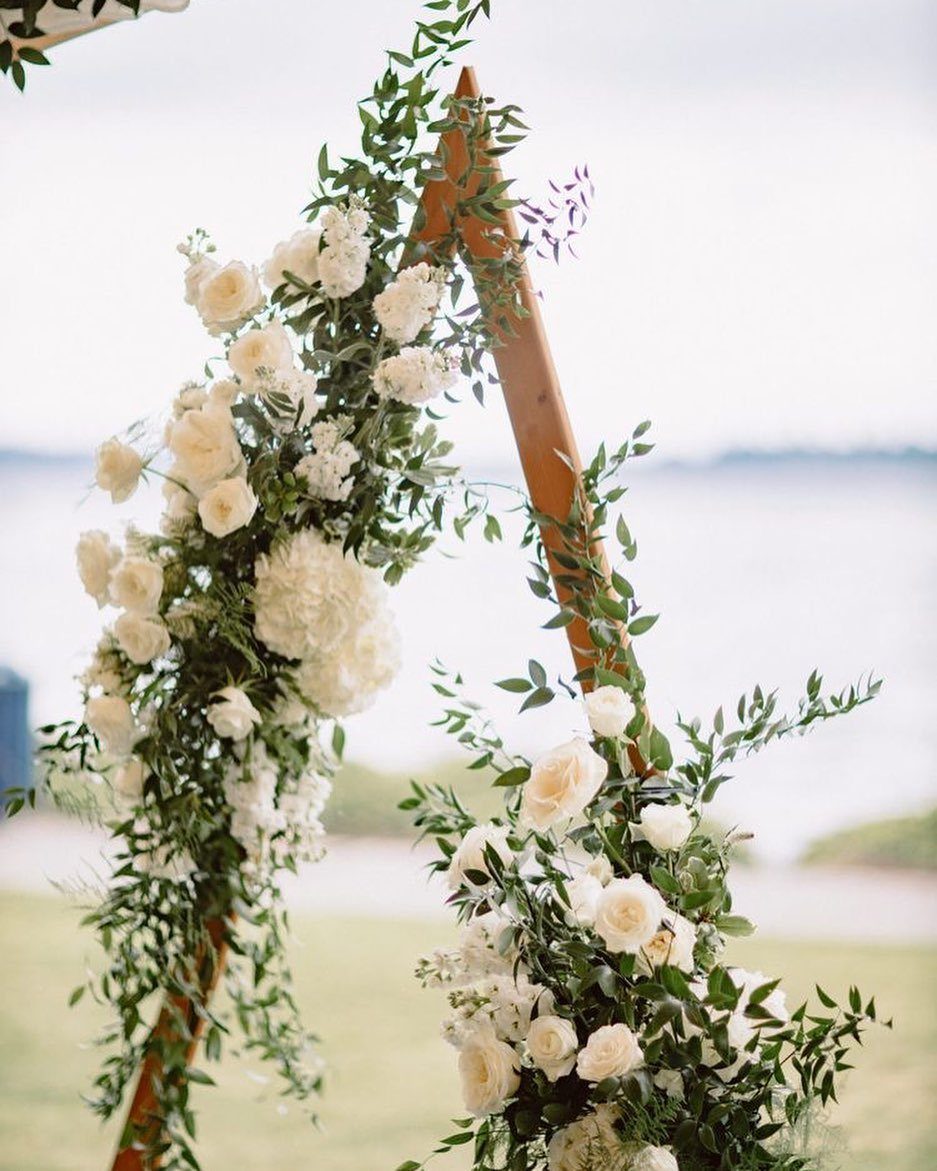 Wooden triangle wedding arch adorned with white roses and lush green foliage outdoors.