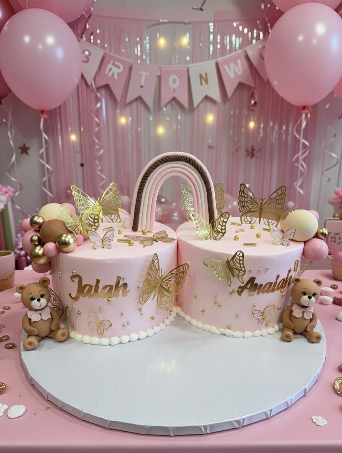Two pink birthday cakes with gold butterflies and a rainbow arch connecting them.