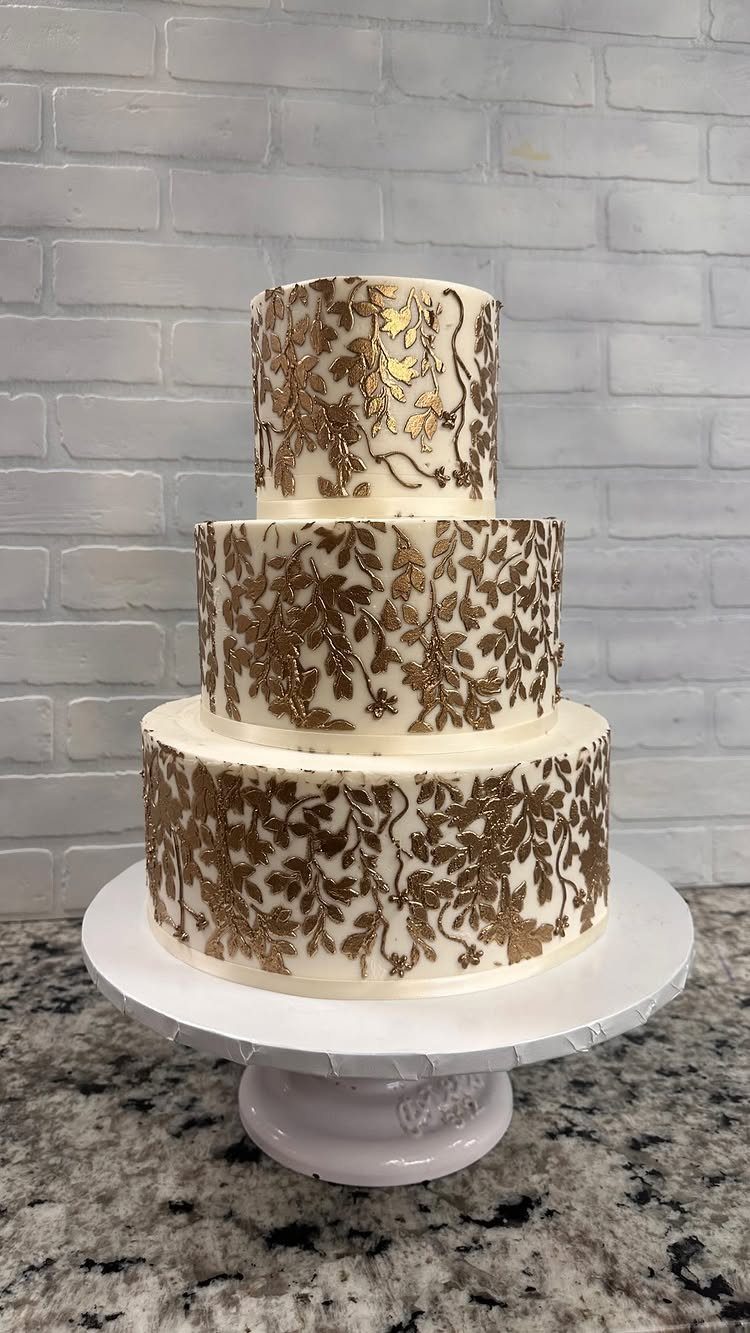 Three-tier white wedding cake with gold leaf floral patterns on a white stand.