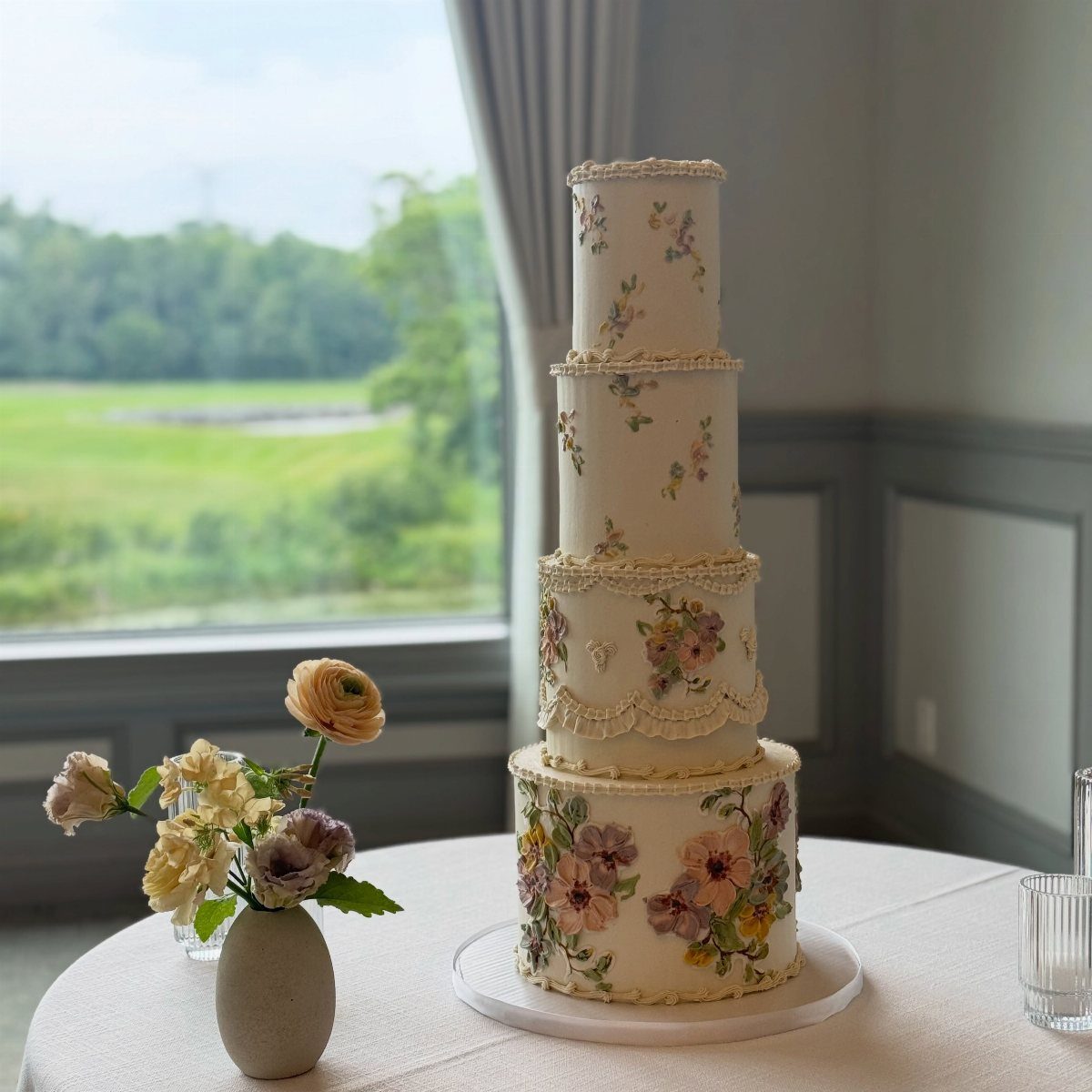 Four-tier white wedding cake with painted impressionistic flowers and delicate piped frosting borders.