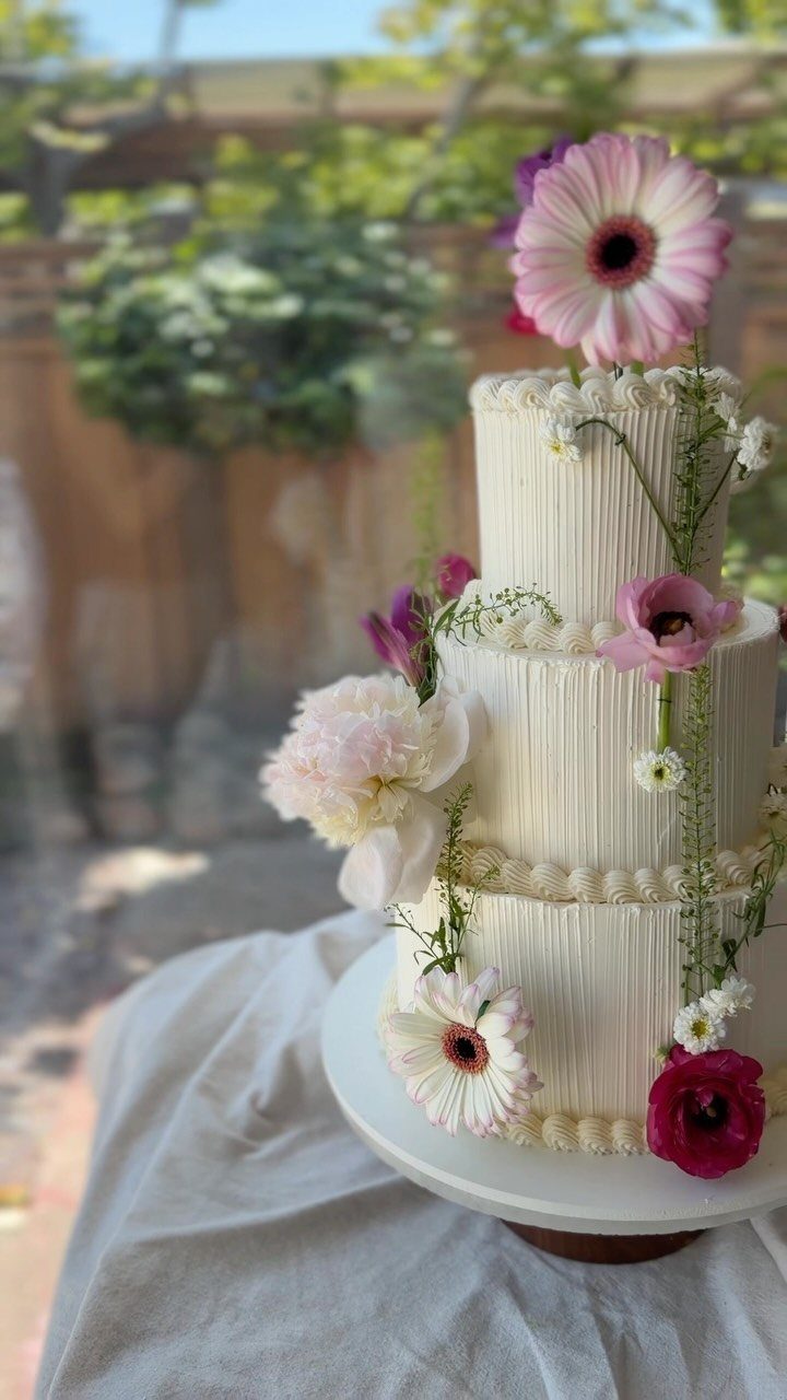 Three-tier ribbed white cake decorated with pink and white wildflowers in a garden.