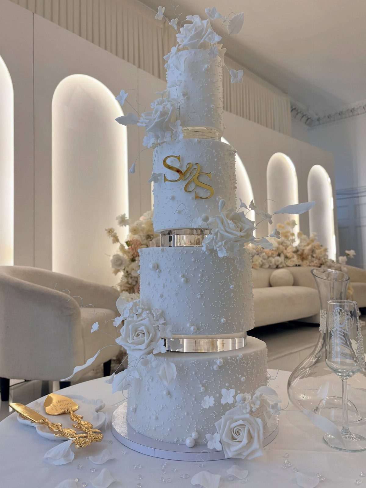 Four-tier white wedding cake with white roses, pearls, and a gold monogram topper.