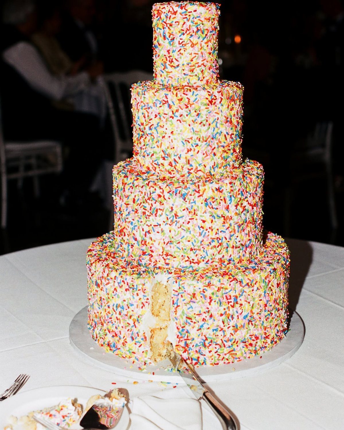 Four-tier wedding cake covered in multicolored rainbow sprinkles on a white table.