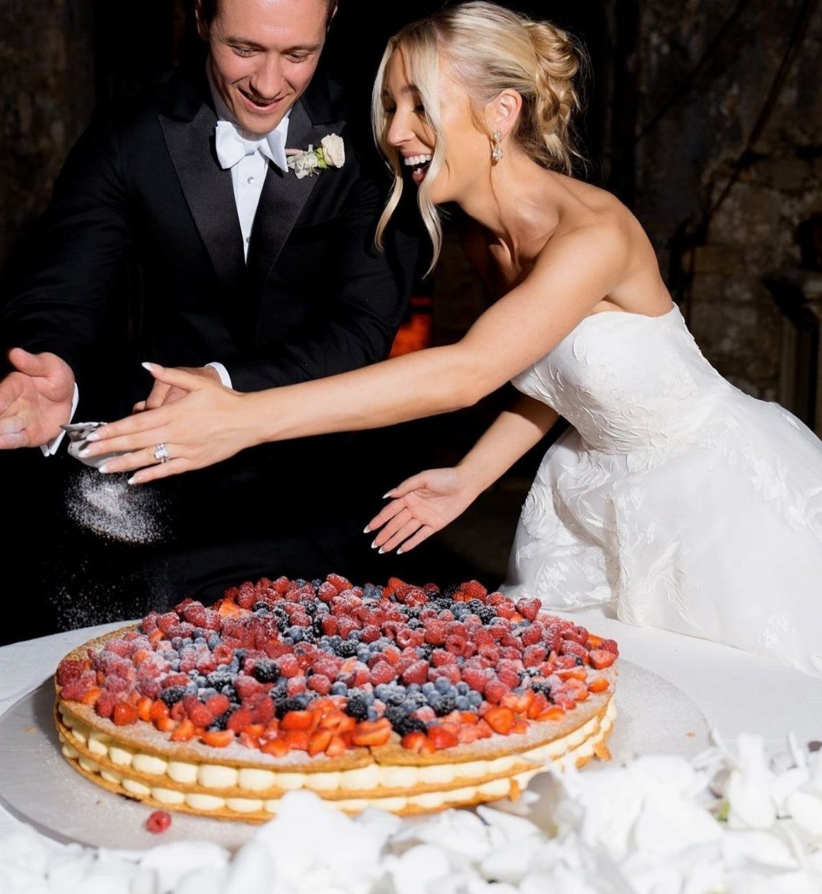 Fruit decorated wedding cake being dusted with powdered sugar by a bride and groom.