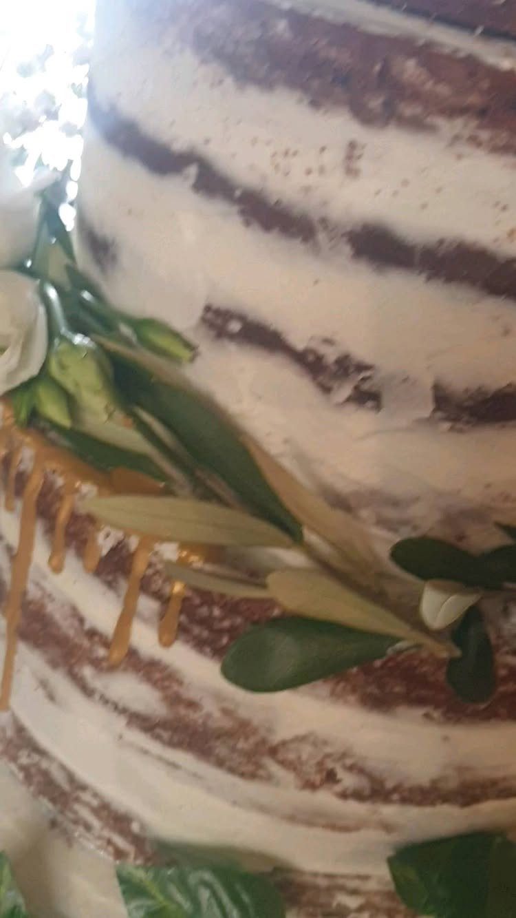 Naked wedding cake with exposed brown layers, white frosting, green leaves, and gold drips.
