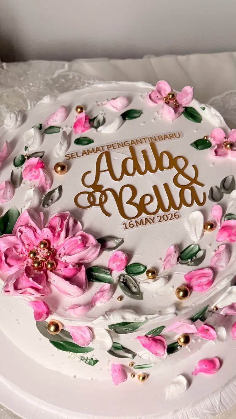 White wedding cake with pink buttercream flowers and a gold personalized name topper.