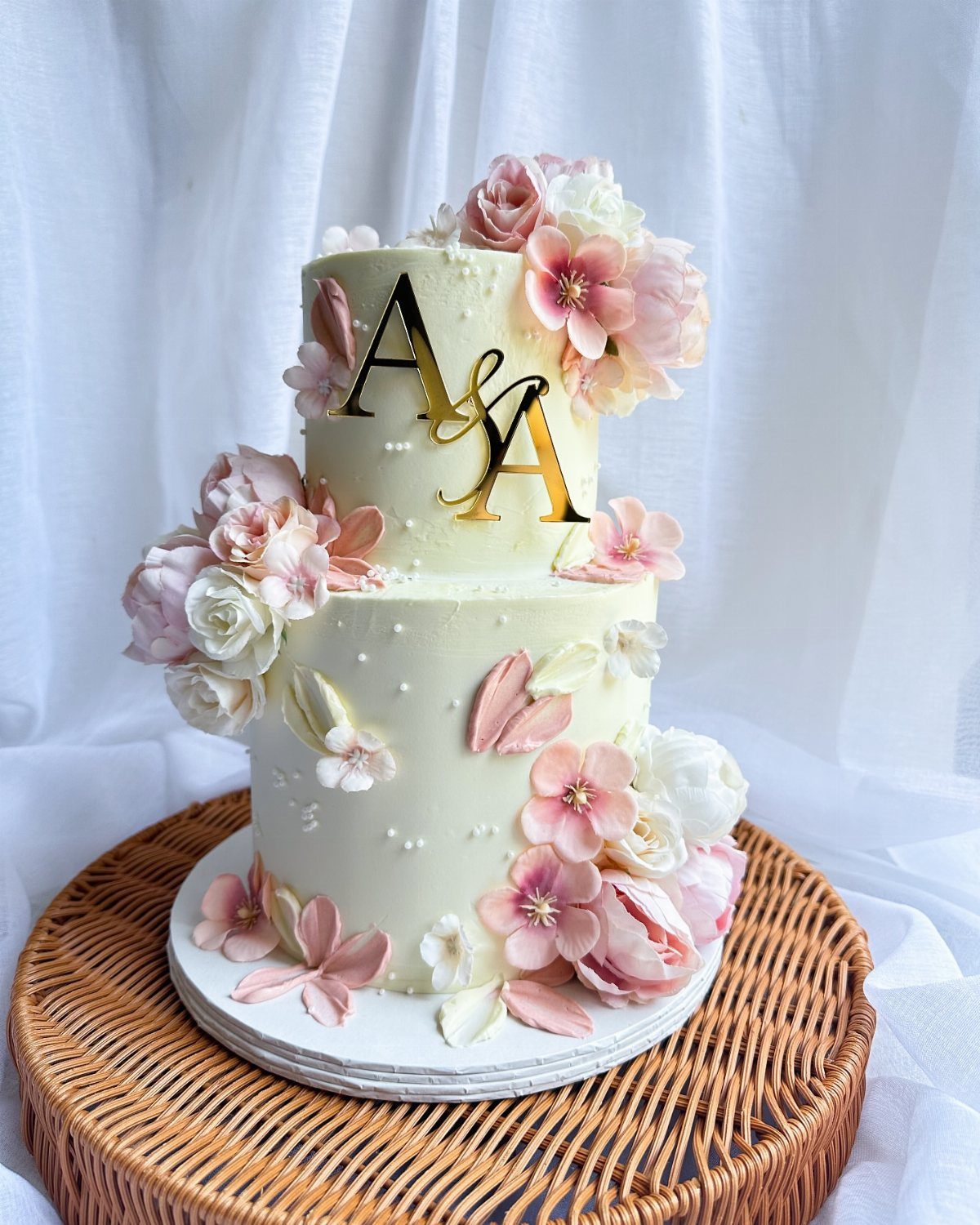 Two-tier white wedding cake with pink flowers and gold A & A initials topper.