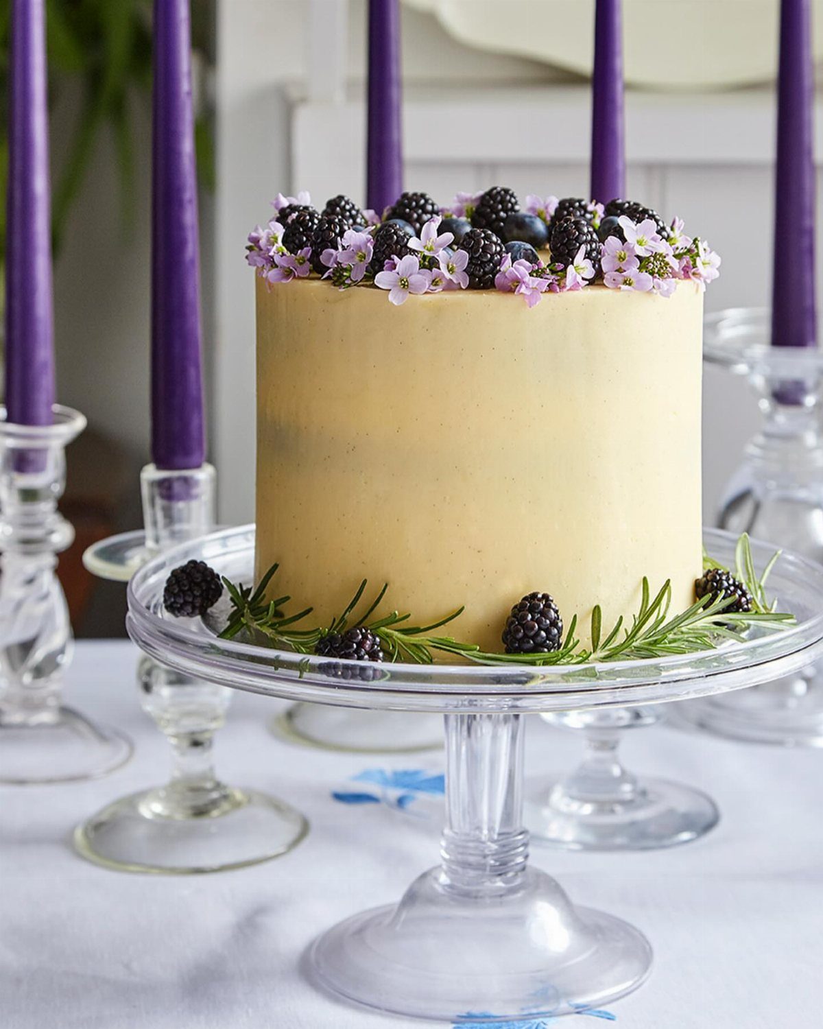 Single-tier yellow cake topped with blackberries and purple flowers on a glass stand.