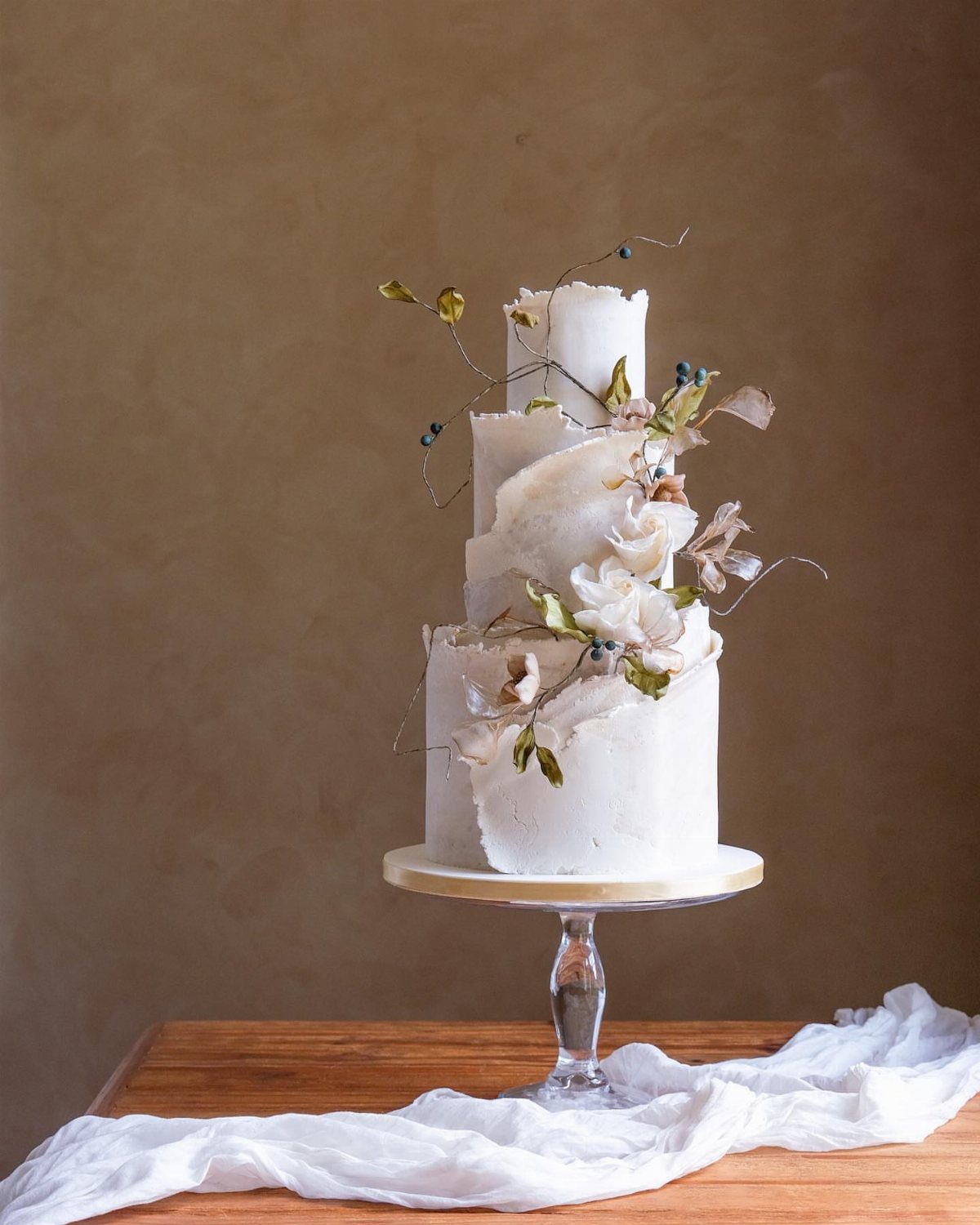 Three-tier white wedding cake with textured frosting and organic floral decorations on a pedestal.