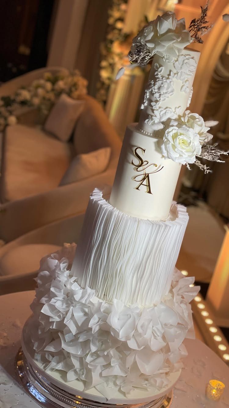 Four-tier white wedding cake with ruffles, pleats, gold initials, and white floral accents.
