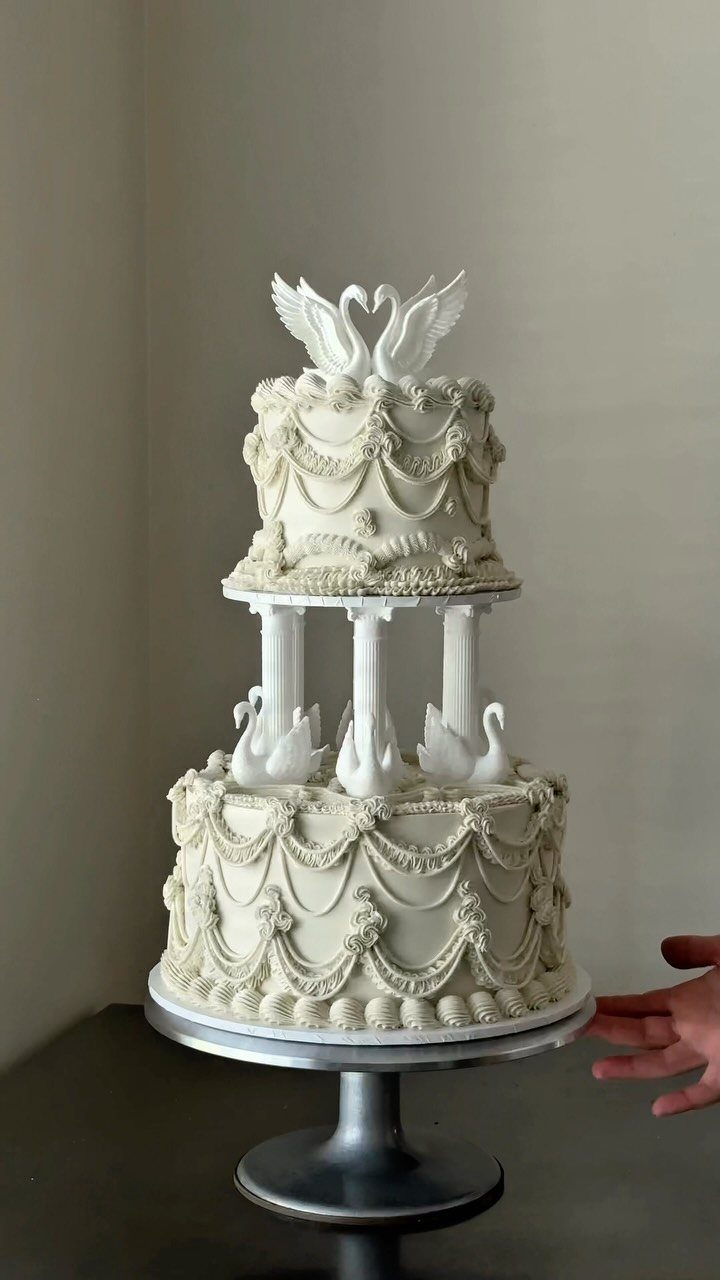 Ornate white wedding cake with vintage buttercream piping and decorative swan figurines.