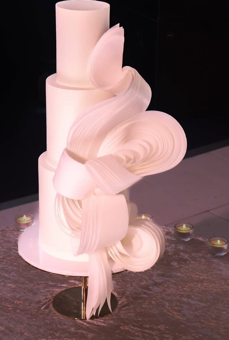 Three-tier white wedding cake with large, swirling white wafer paper ribbons and candles.