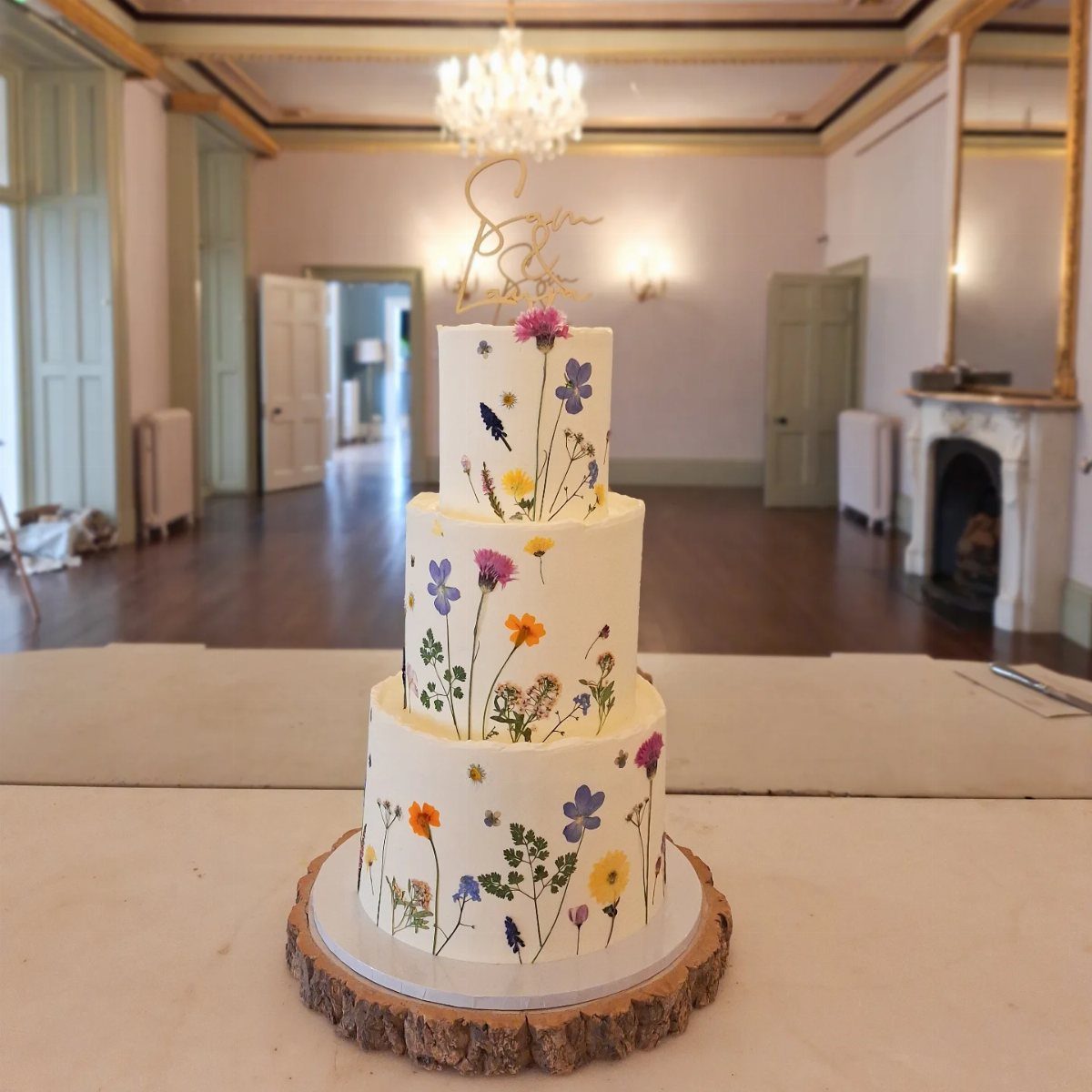 37 Wedding Cakes with Flowers to Inspire Your Design 25 Three-tier white wedding cake decorated with colorful pressed edible flowers on a wooden base.