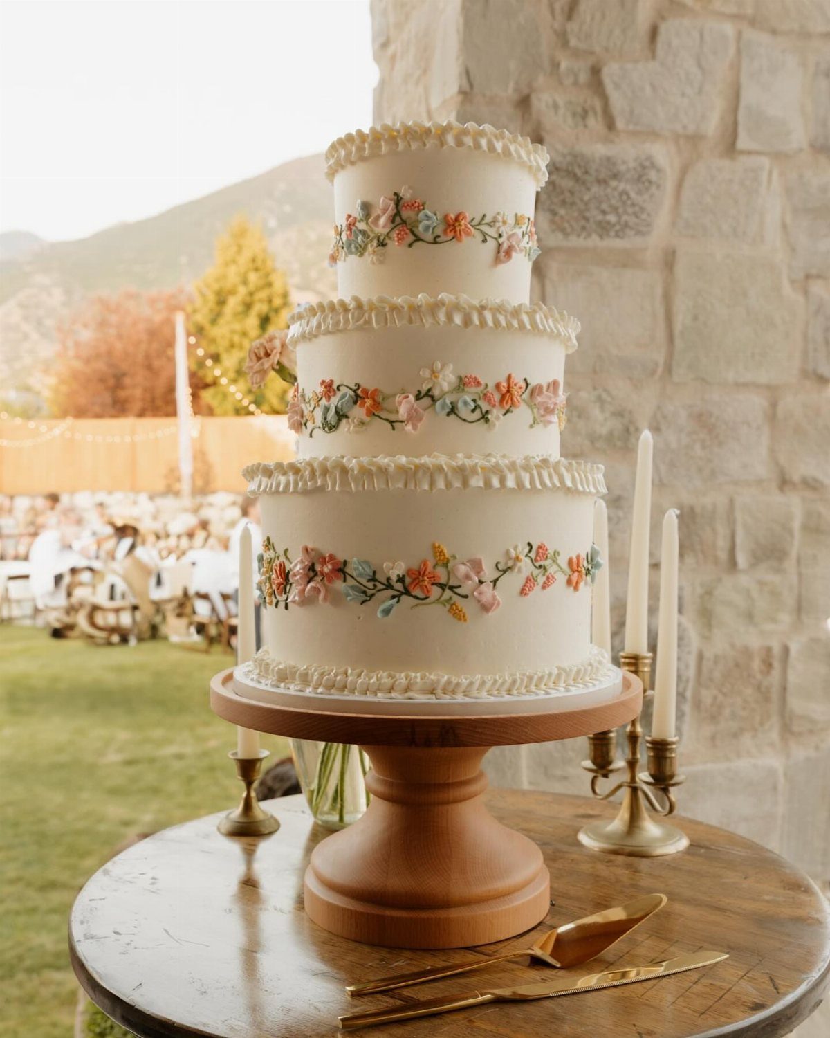 37 Wedding Cakes with Flowers to Inspire Your Design 29 Three-tier white wedding cake with colorful floral bands on a wooden pedestal.