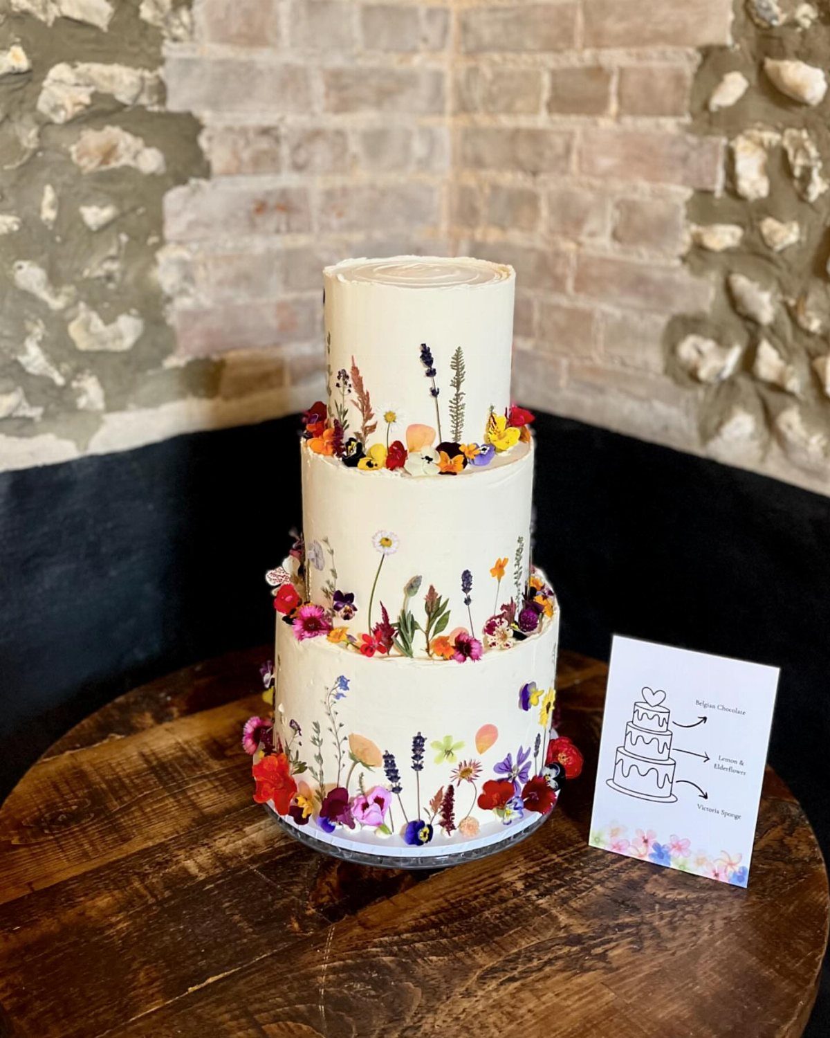 37 Wedding Cakes with Flowers to Inspire Your Design 10 Three-tier white cake decorated with pressed and natural colorful wildflowers on a wooden table.