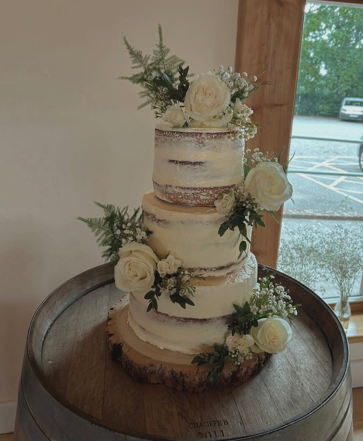 37 Wedding Cakes with Flowers to Inspire Your Design 14 Three-tier semi-naked wedding cake with white roses and greenery on a wooden barrel.