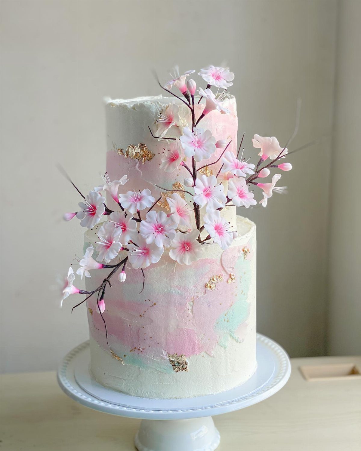 37 Wedding Cakes with Flowers to Inspire Your Design 19 Two-tier white cake decorated with pink cherry blossoms, gold leaf, and watercolor frosting.
