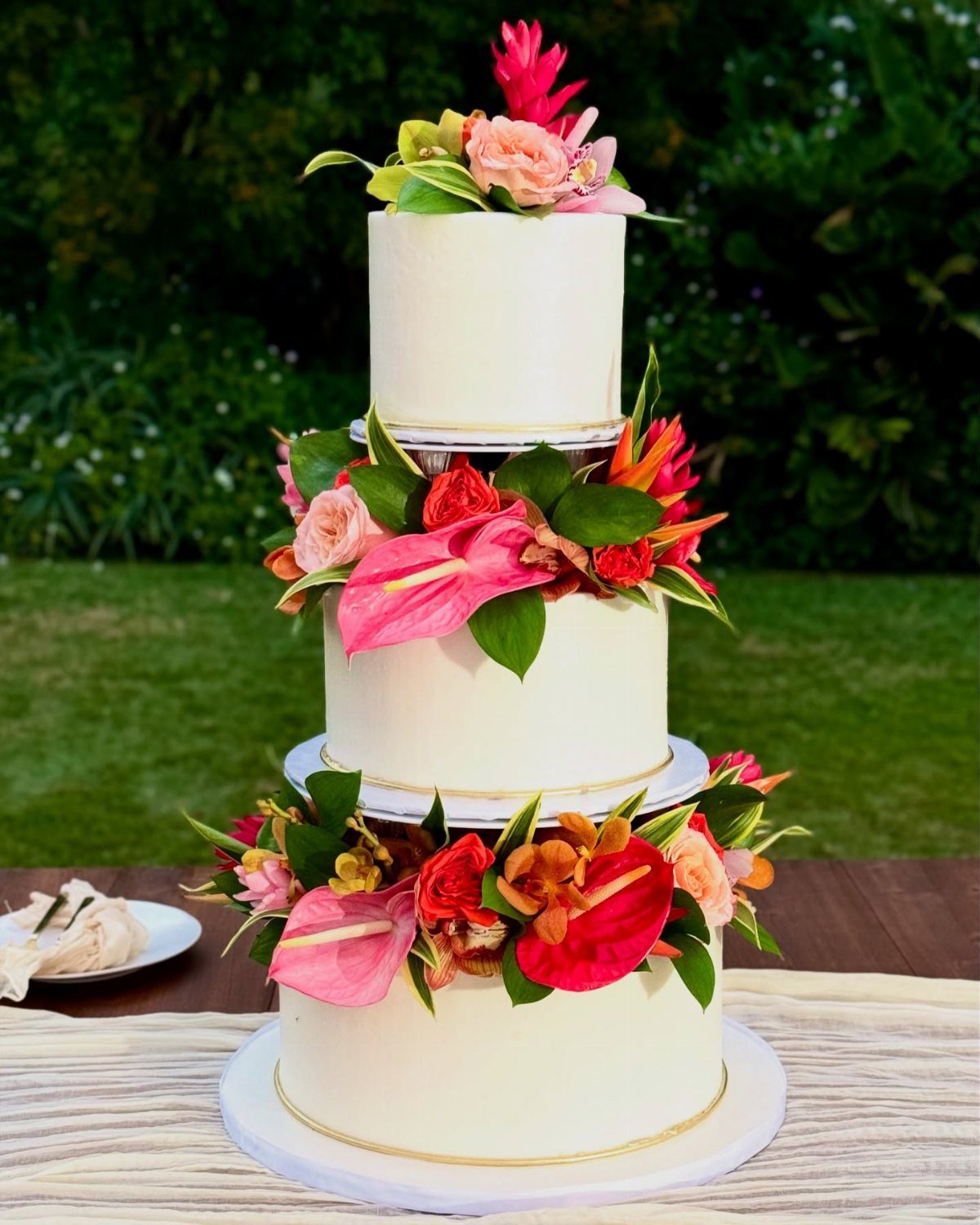 37 Wedding Cakes with Flowers to Inspire Your Design 17 Three-tier white wedding cake adorned with bright pink anthuriums and tropical greenery outdoors.