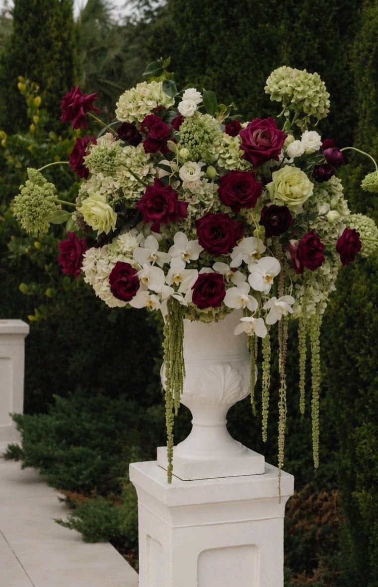 Burgundy roses and green hydrangeas in a white urn against a garden backdrop.