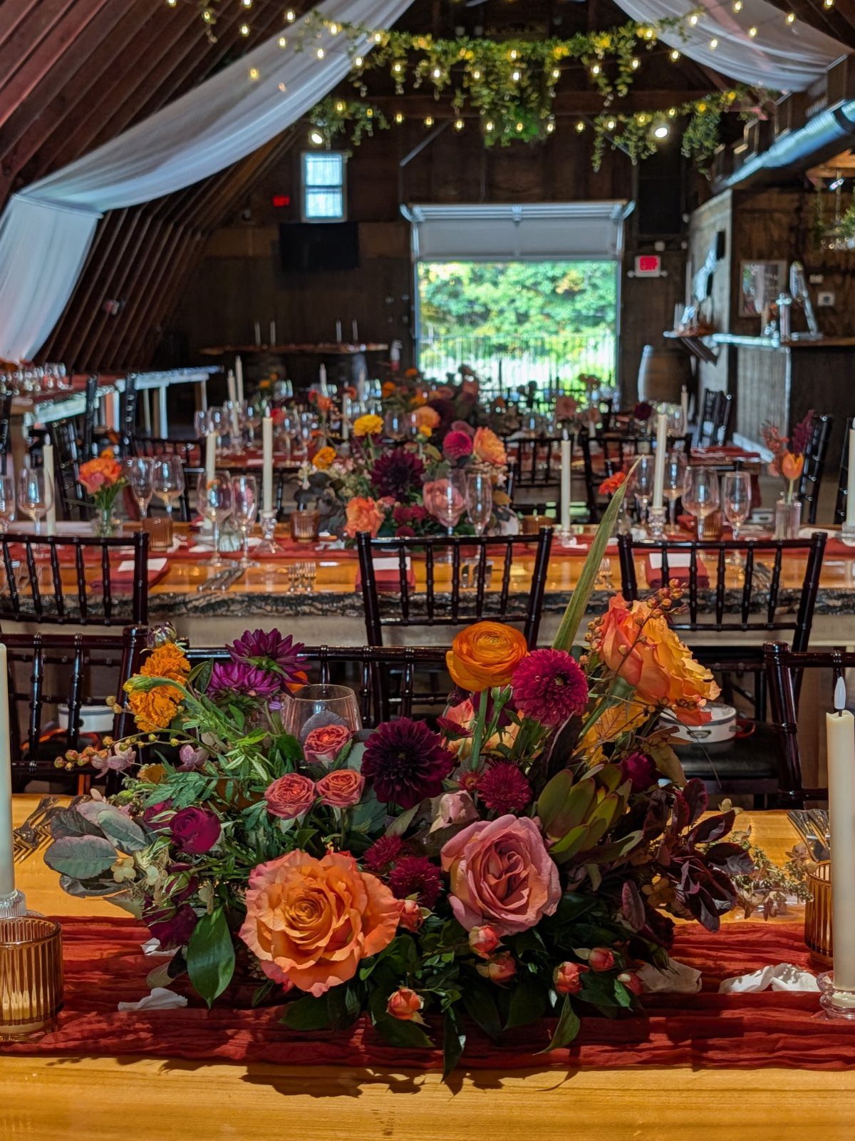 Burgundy and orange wedding flower centerpieces on wooden tables in a rustic barn.