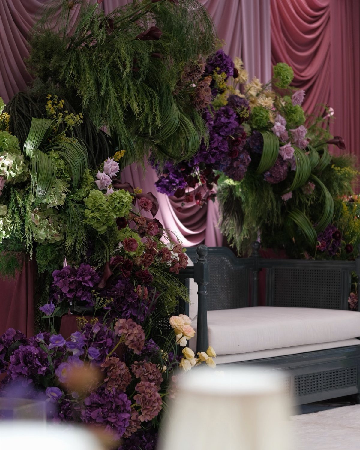Deep purple flowers and green foliage arrangement against draped mauve fabric curtains.
