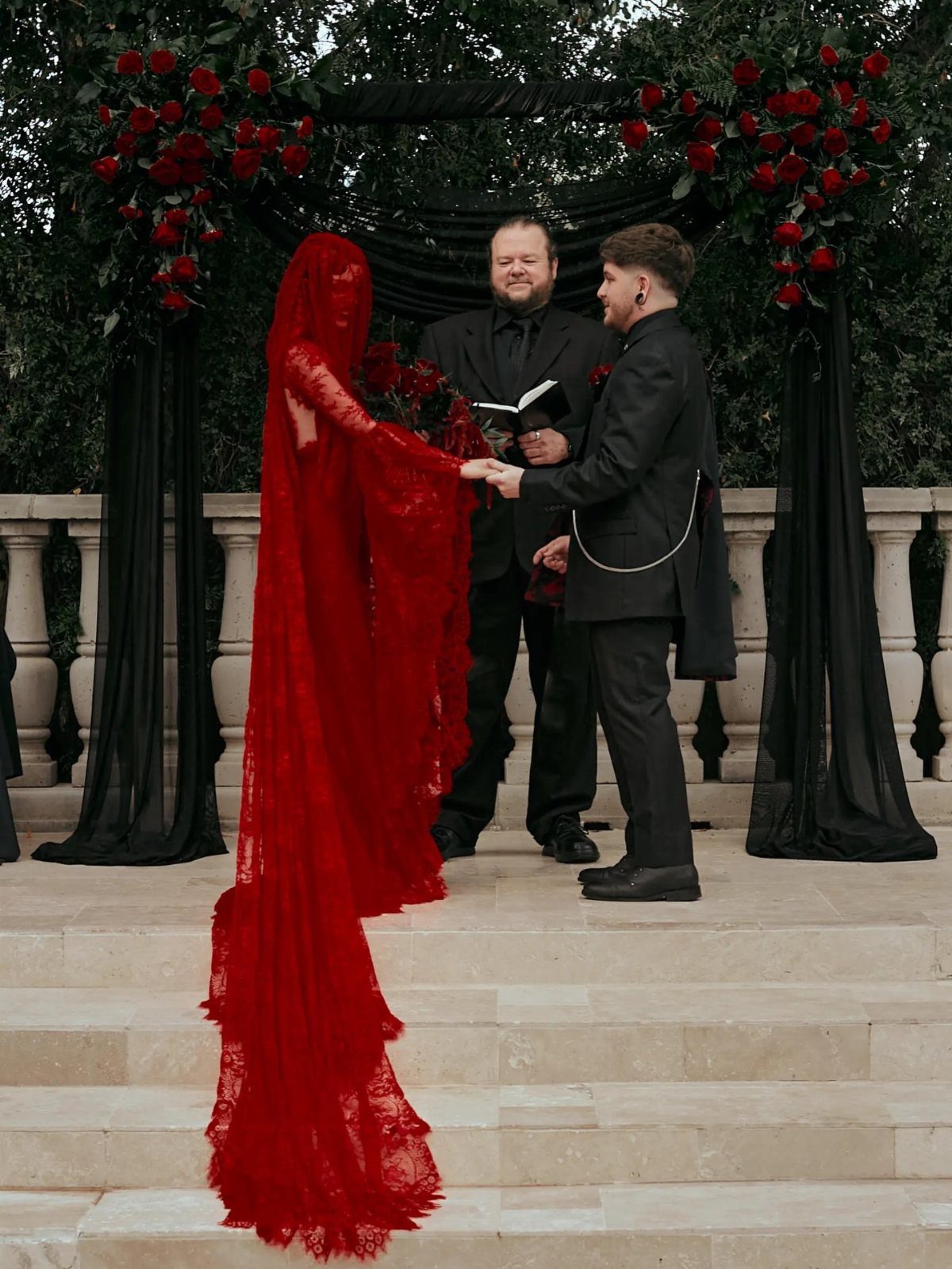 Bride in red lace gown and groom in black suit at outdoor wedding ceremony