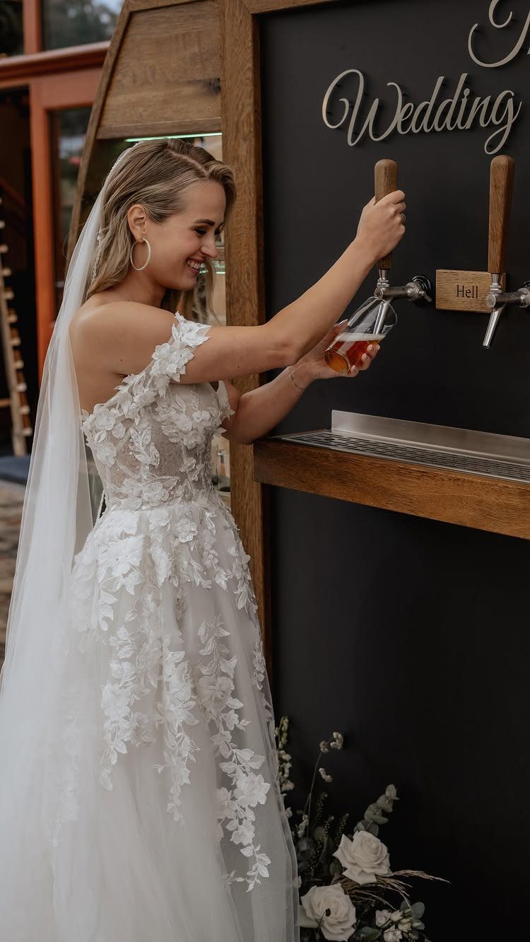 Bride in a white lace gown pouring beer from a brewery tap system.