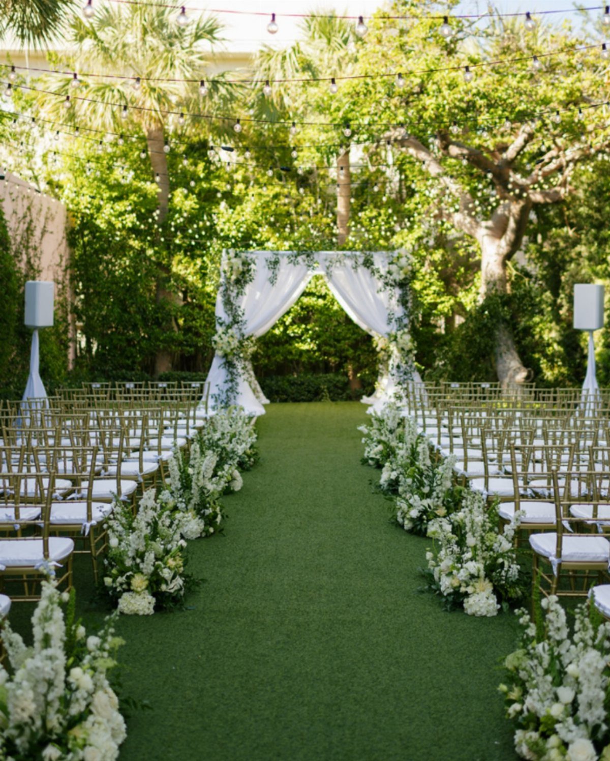 Outdoor wedding ceremony setup with white floral arch and gold chairs in a garden