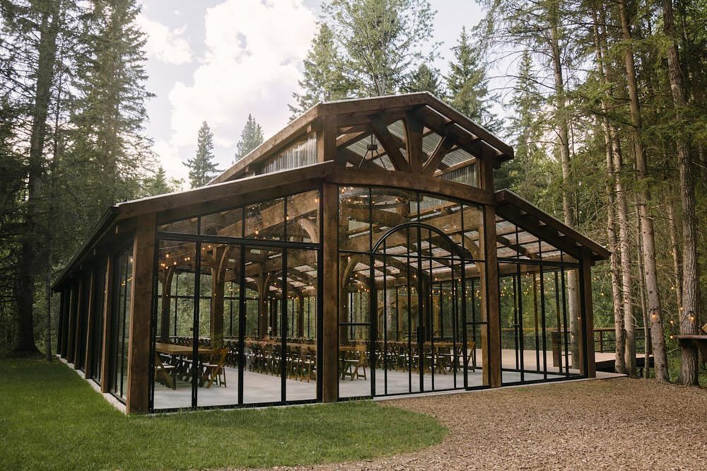 Glass-walled timber pavilion with dining tables set inside a dense evergreen forest.