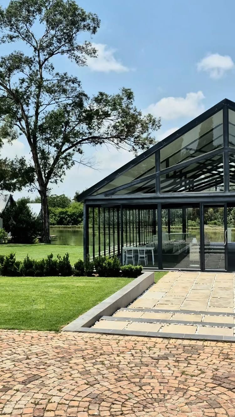 Modern glass pavilion with a long dining table overlooking a pond and green lawn.