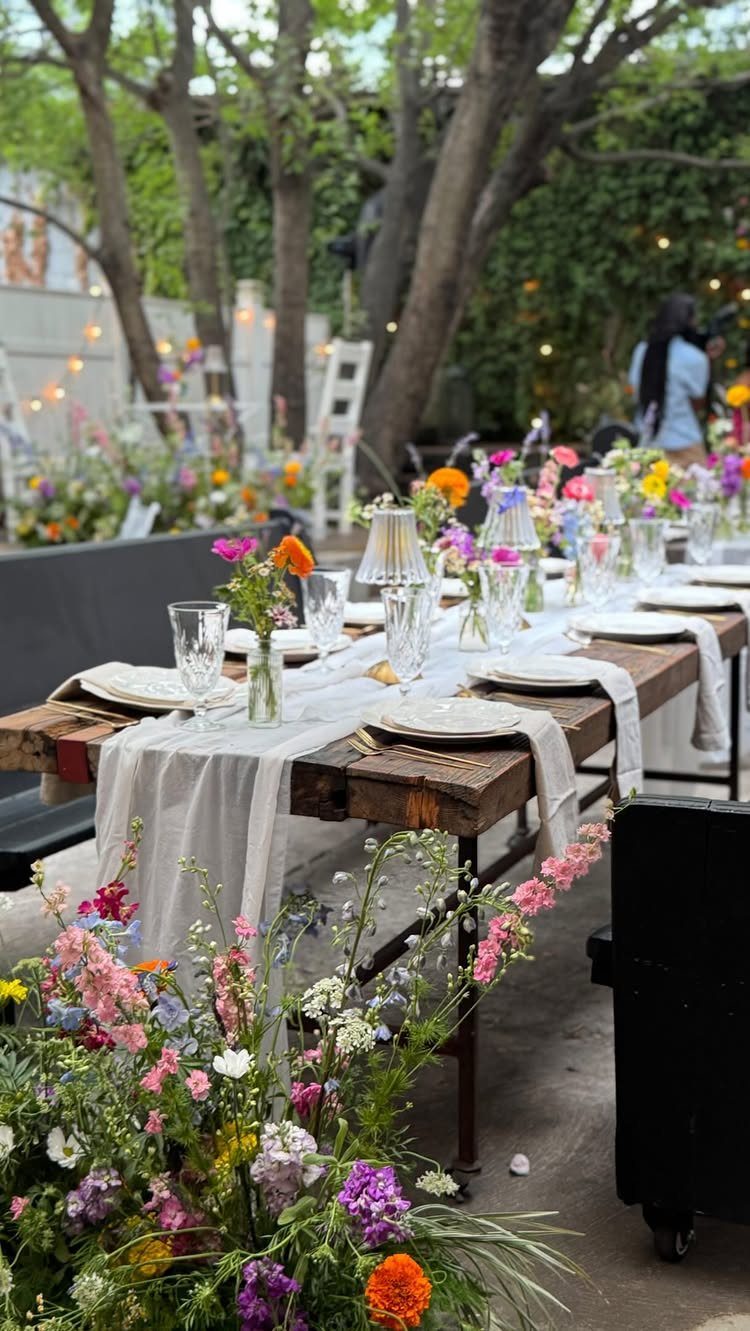 Wooden dining table with white linens and colorful wildflowers set outdoors under trees.