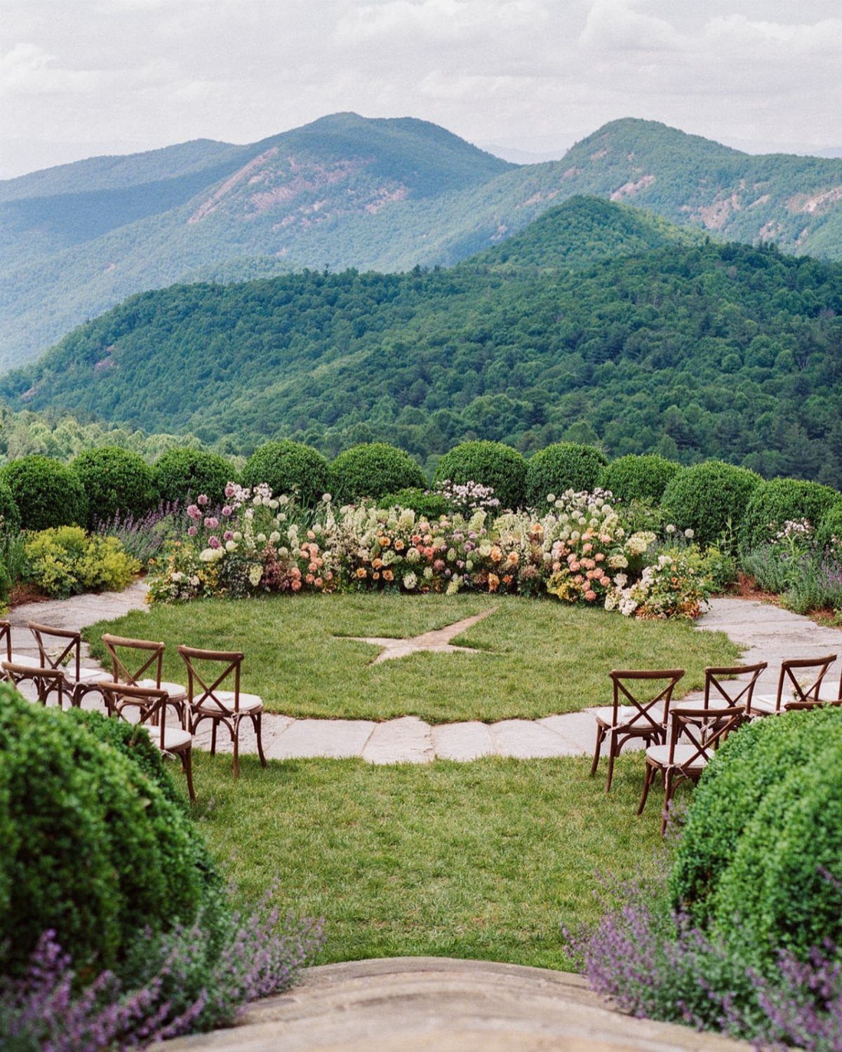 Outdoor wedding ceremony setup with wooden chairs and flowers overlooking green mountains.