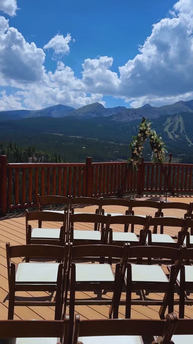 Wooden wedding chairs and floral arch on a deck overlooking distant blue mountains.