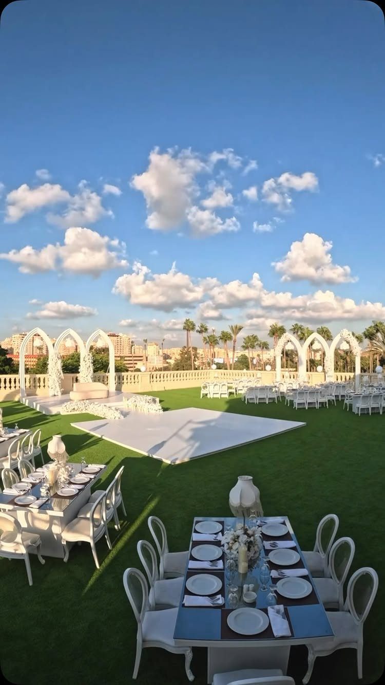 Rooftop wedding setup with white tables, chairs, and decorative arches on green turf.