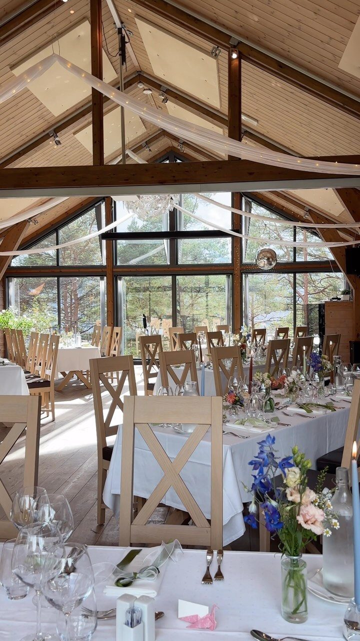 Wedding reception tables with white linens and wooden chairs in a sunlit timber hall.