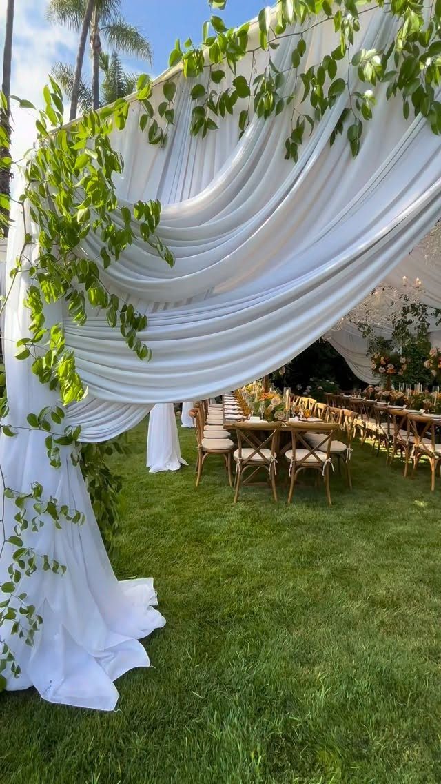 White draped fabric canopy with greenery over wooden dining tables on a lawn
