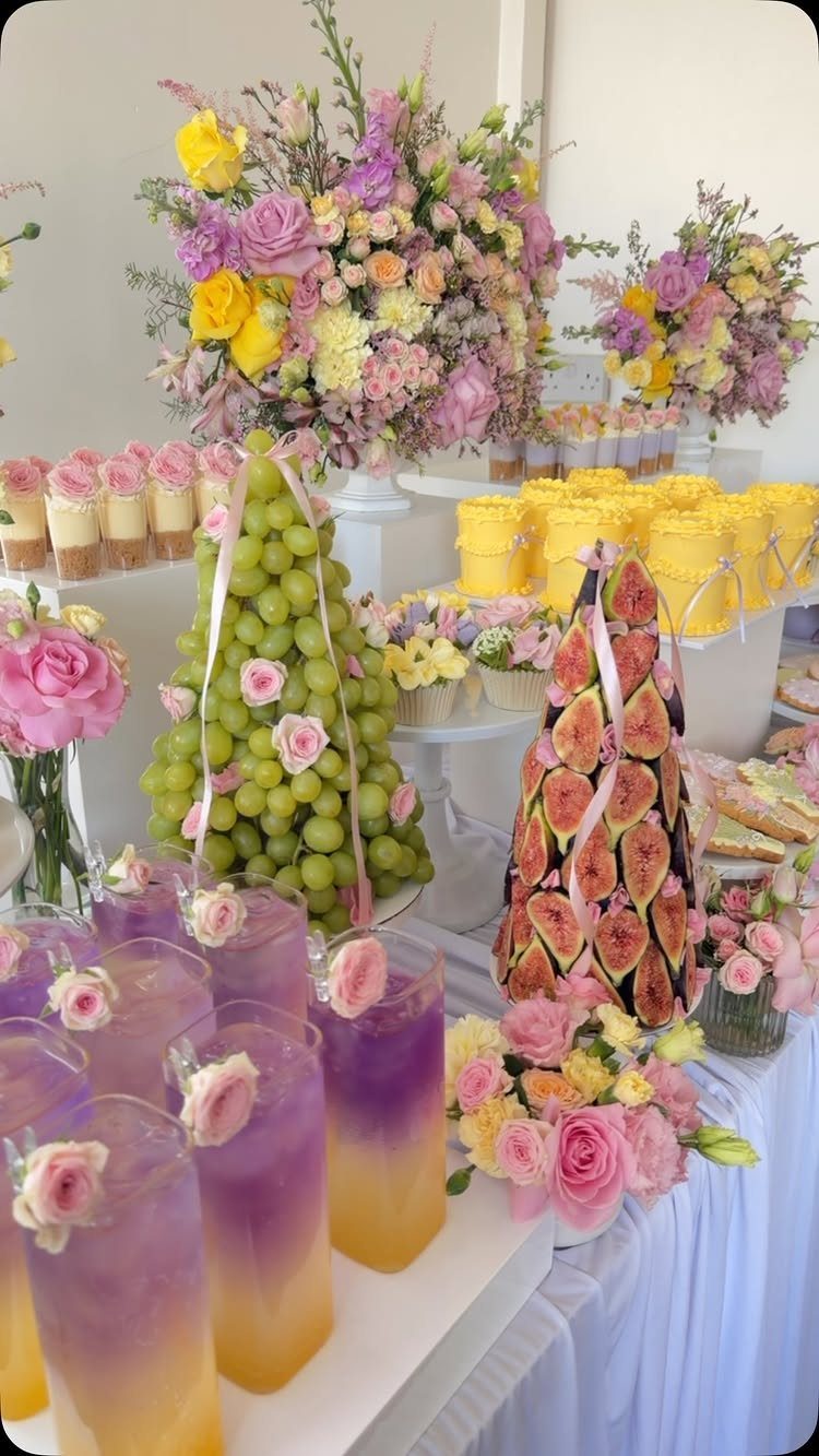 Dessert grazing table with fruit towers, colorful drinks, and pink and yellow floral arrangements.