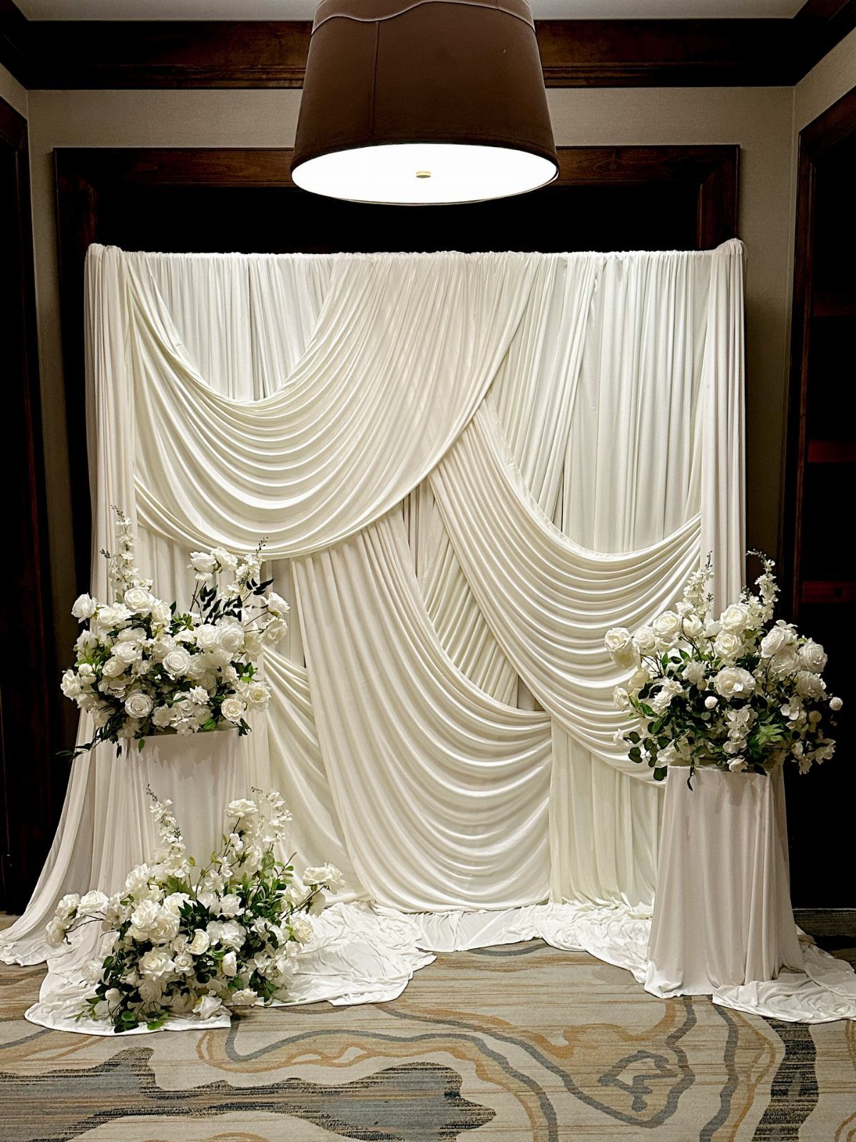 Wedding Mood Board Ideas: 39 Styles from Celestial to Coastal 38 White draped fabric backdrop with white floral arrangements on pedestals in a room.