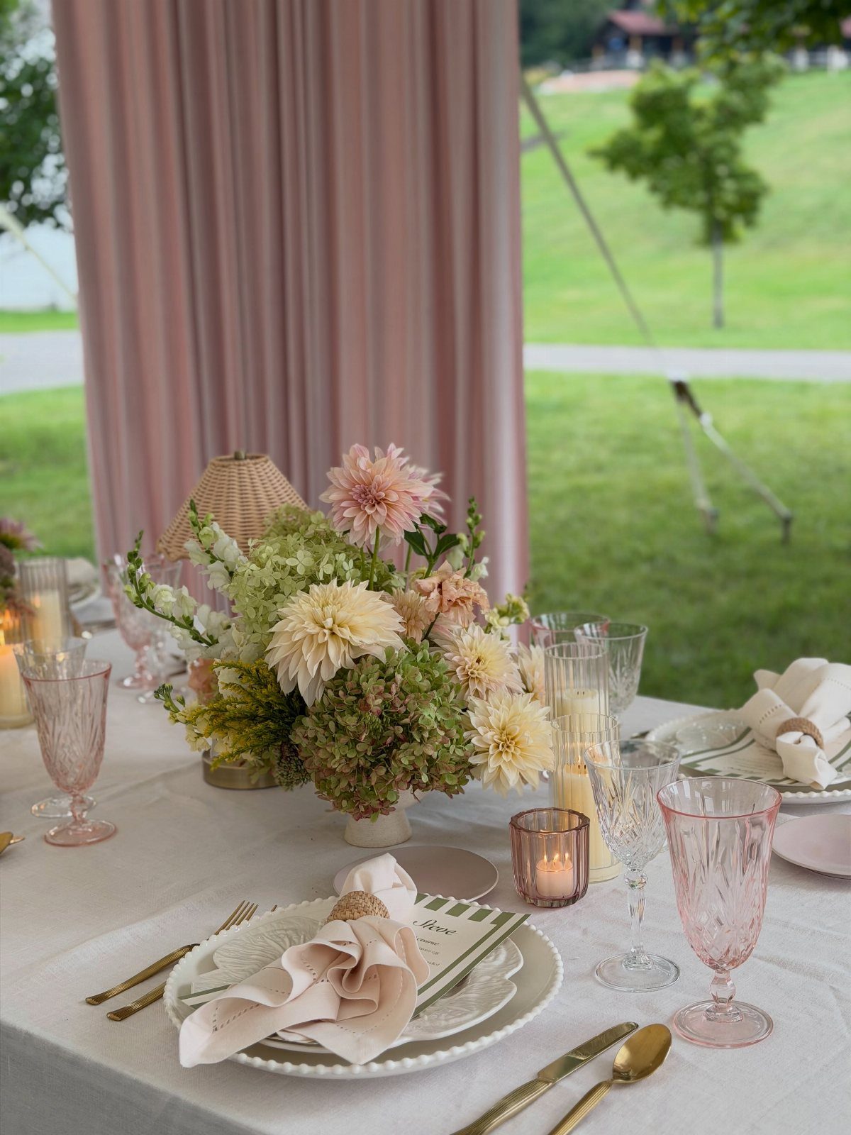 Wedding table setting with pink glassware and floral centerpiece against pink curtains.