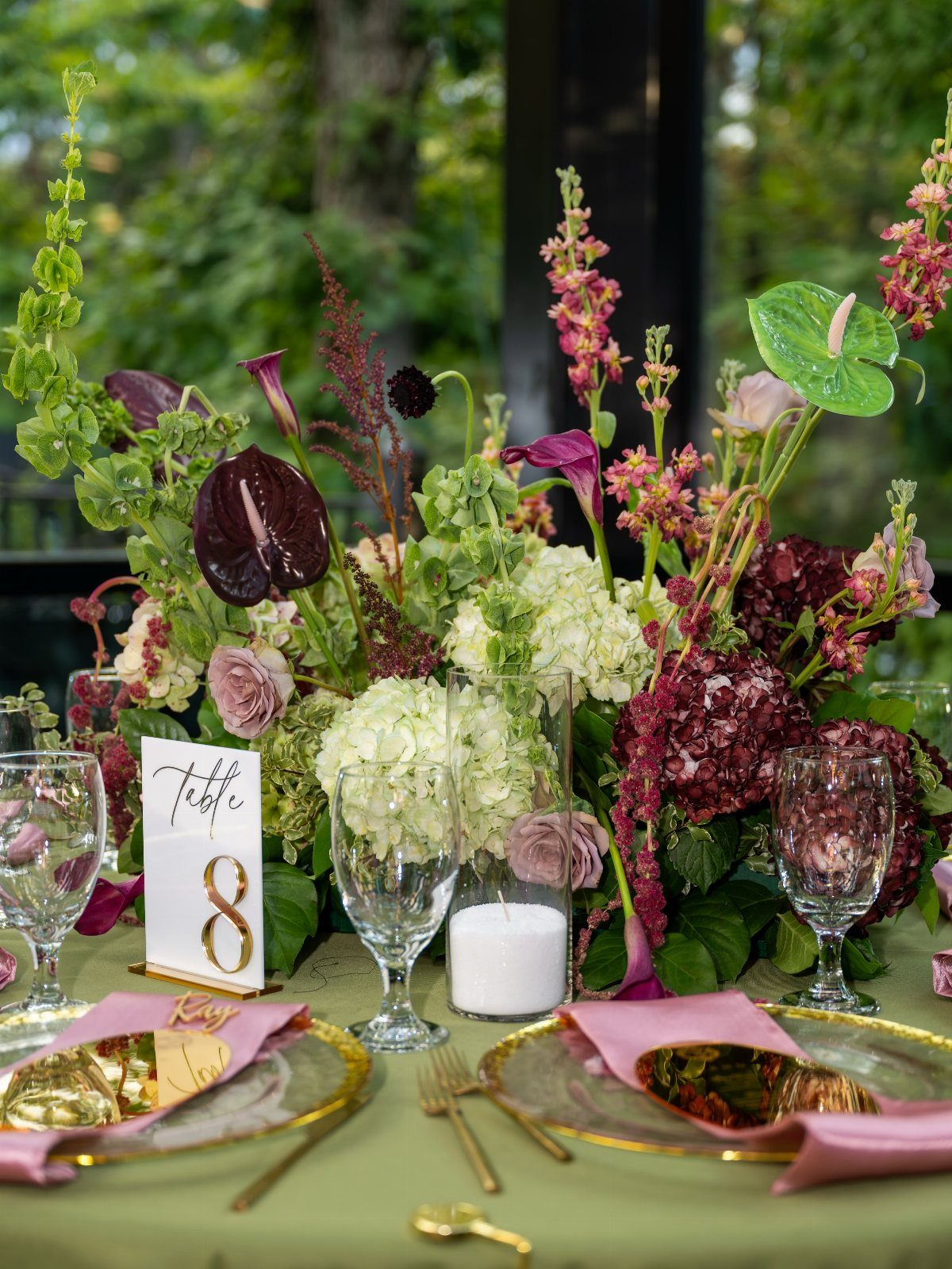 Wedding table setting with green cloth, gold plates, and burgundy and white flowers.