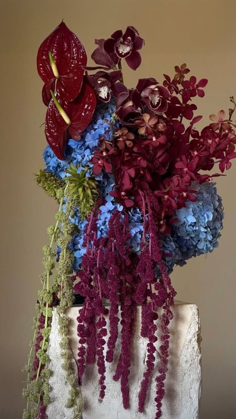 Burgundy and blue floral arrangement on a white stone pedestal against a beige wall.