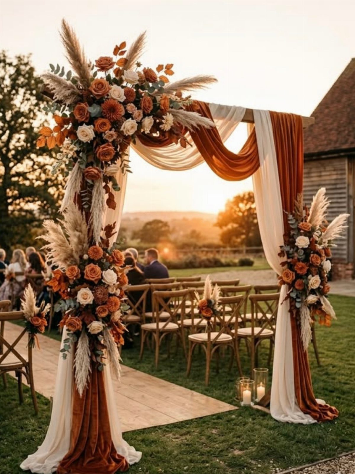 Wedding arch with orange and cream fabrics, pampas grass, and autumn-colored roses at sunset
